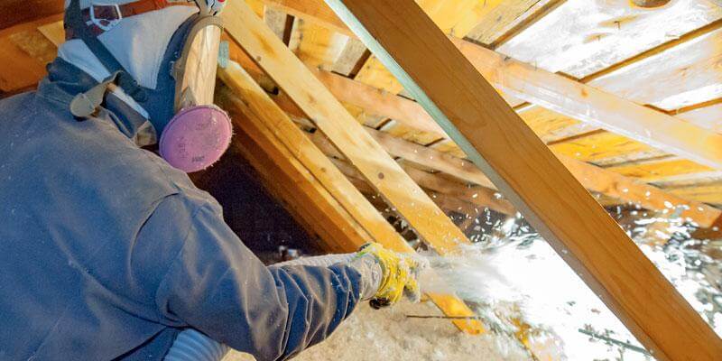 person spraying insulation into attic while wearing mask